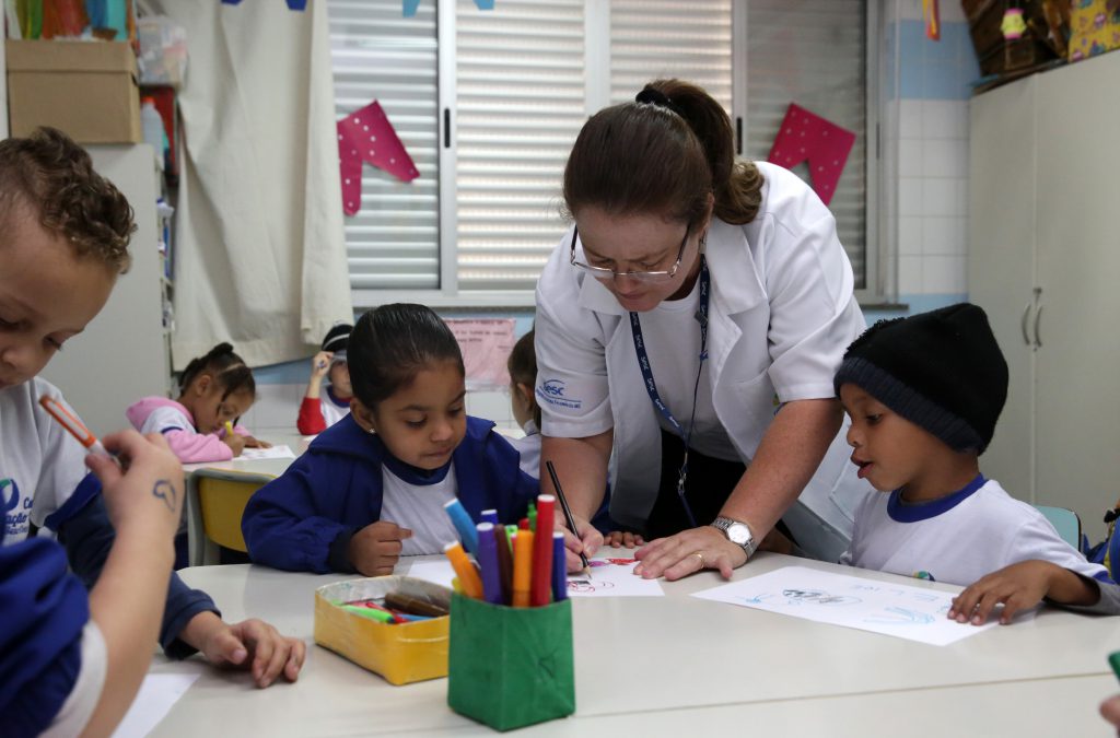 duas crianças pequenas observam professora orientar atividades didáticas em uma emsa